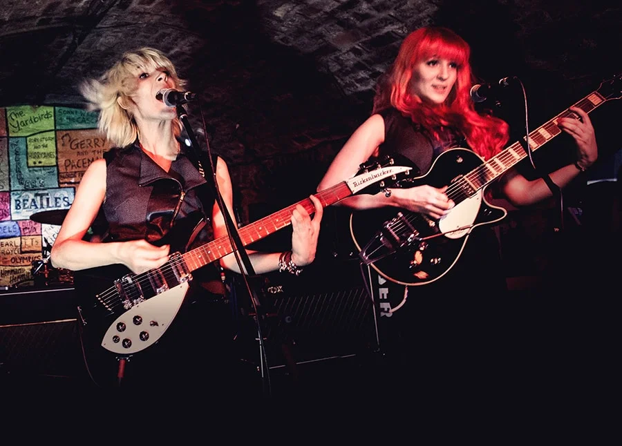 Mona and Lisa with guitars in front of the colorful backdrop of the world-known vaulted front stage of the Liverpool Cavern Club